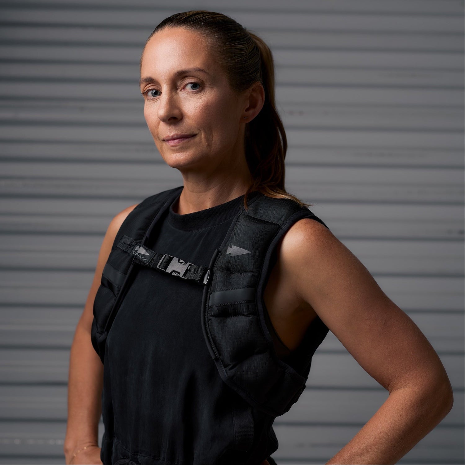 A confident woman in a black sleeveless top wearing a weighted vest stands against a gray striped background.