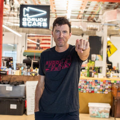 A man wearing the Buddy FCKR Friday Tee - Tri-BlendX stands indoors, pointing at the camera with a “GORUCK SCARS” sign behind him—showcasing his GORUCK pride and readiness to pay it forward.