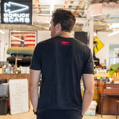 A man stands indoors, facing away, wearing the “It’s Not About You - Tri-BlendX - Unisex” t-shirt in black—crafted from premium Tri-BlendX fabric and printed in the USA—with equipment and signs visible in the background.