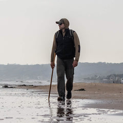 A person strolls along the beach with a walking stick, clad in a hooded jacket and sunglasses. Their GORUCK Performance TAC Hat - TOUGHDRY, made from sweat-wicking material, keeps them comfortable under the overcast sky. Distant hills and buildings fade into the background.