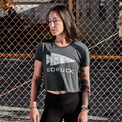 A person with long hair stands in front of a chain-link fence, wearing a Women's Cropped Tee - Poly-Blend by GORUCK in gray and black leggings. The individual has tattoos on their arms and gazes to the side under bright sunlight.