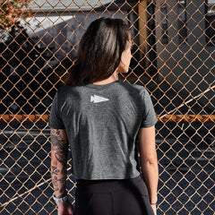 A person with long dark hair faces a chain-link fence, wearing the Women's Cropped Tee - Poly-Blend by GORUCK. The gray top features a triangular symbol on the back and highlights their tattooed forearm, as sunlight creates shadows on the fence and ground.