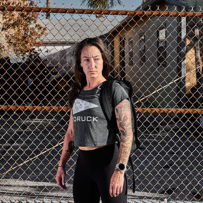 A person with long hair stands in front of a chain-link fence, wearing a rugged gray Women's Cropped Tee by GORUCK and black leggings. They're carrying a backpack, their tattooed arms catching the sunlight as they enjoy the sunny day outdoors, embodying style and resilience.