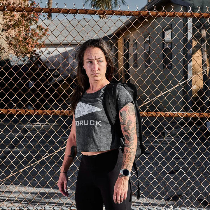 A person with long hair stands in front of a chain-link fence, wearing a rugged gray Women's Cropped Tee by GORUCK and black leggings. They're carrying a backpack, their tattooed arms catching the sunlight as they enjoy the sunny day outdoors, embodying style and resilience.