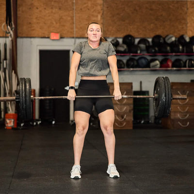 Dressed in a premium GORUCK Women's Subtle Spearhead x CrossFit Cropped Tee, a poly-blend, and black shorts, a person lifts a barbell in the gym. The setting features essential workout gear like medicine balls and weights, creating an ideal environment for fitness enthusiasts.