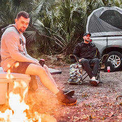 Two men relaxing by a campfire with backpack, camping chair, and truck with rooftop tent in forest setting