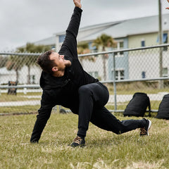 Man in black GORUCK fleece jacket and pants stretching outdoors on grass near chain-link fence with backpacks