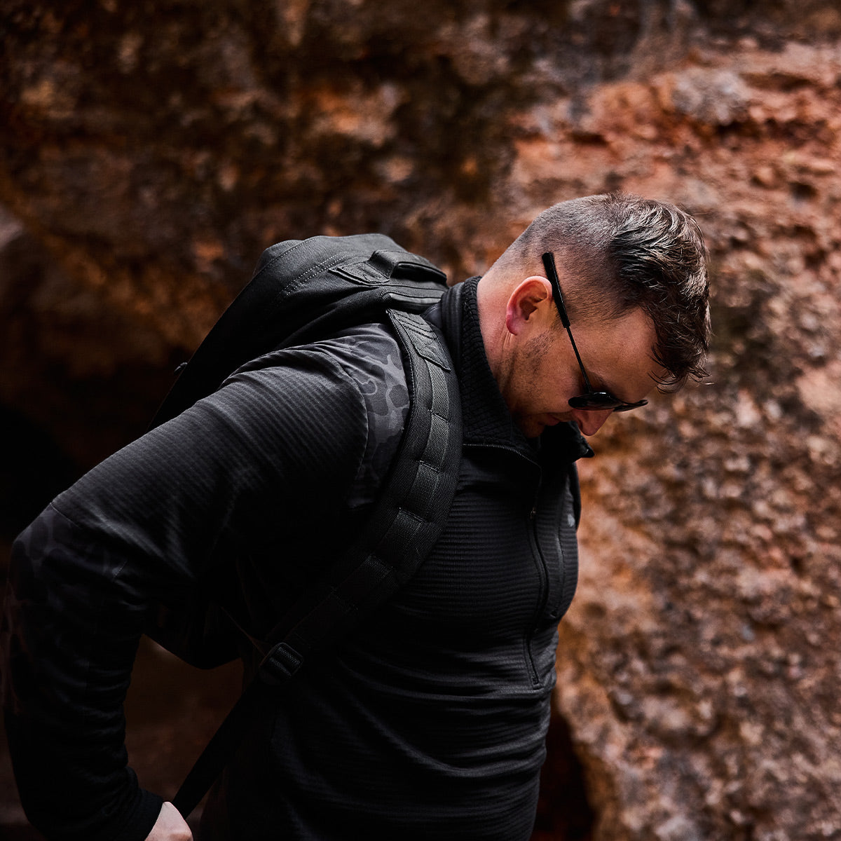 A man in a sleek black Men’s Half Zip - Polartec Grid Fleece and sunglasses stands outdoors, his ToughDry backpack slung over his shoulder against a rugged rocky backdrop.
