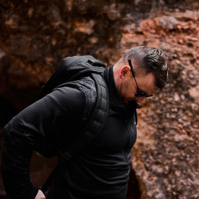 A man in a sleek black Men’s Half Zip - Polartec Grid Fleece and sunglasses stands outdoors, his ToughDry backpack slung over his shoulder against a rugged rocky backdrop.