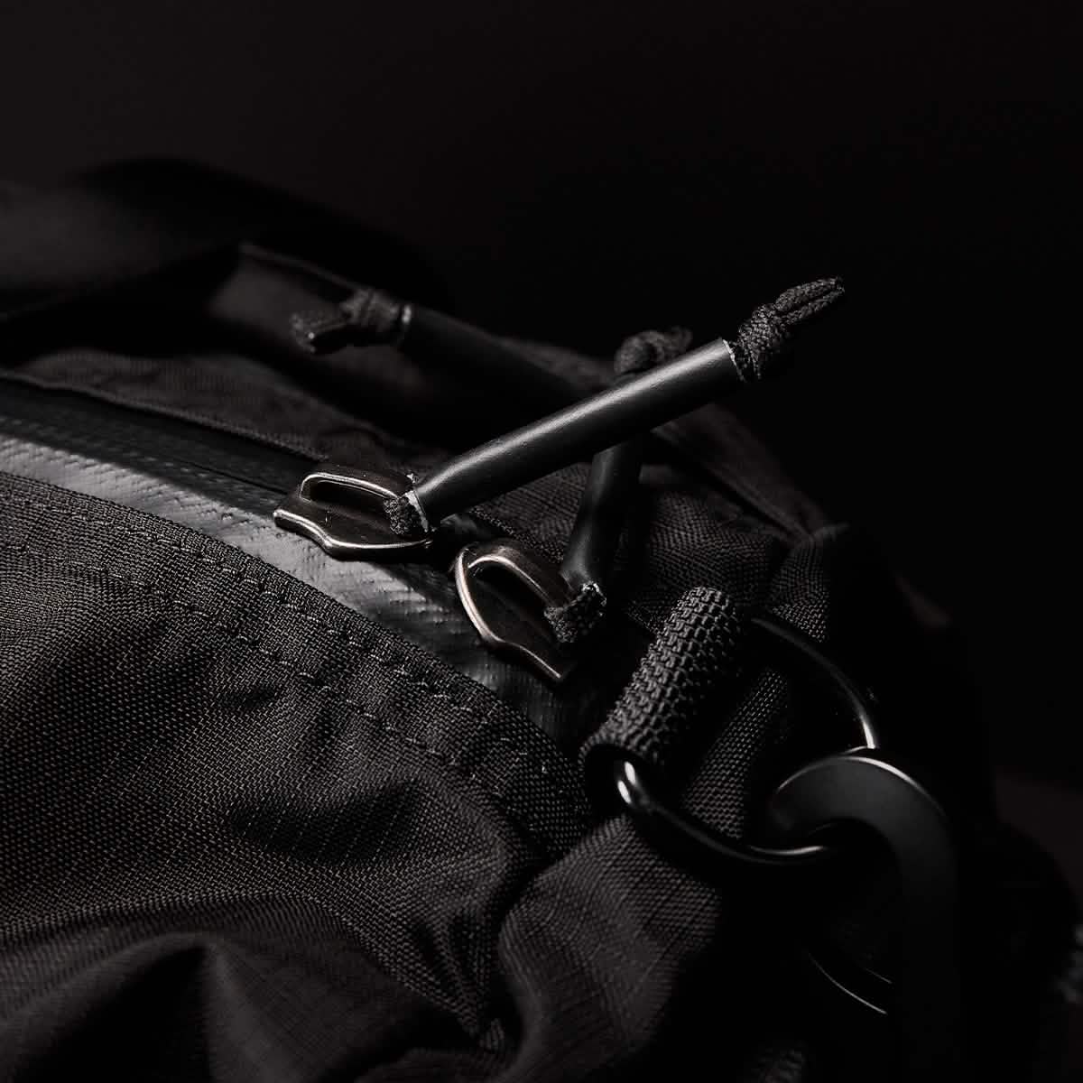 Close-up of a GORUCK Gym Bag - Ripstop ROBIC® backpack highlights the zipper and pull tags. The textured ripstop nylon with detailed stitching features metal zippers and loops. The dark background emphasizes the bags design, suggesting a concealed shoe compartment for extra convenience.