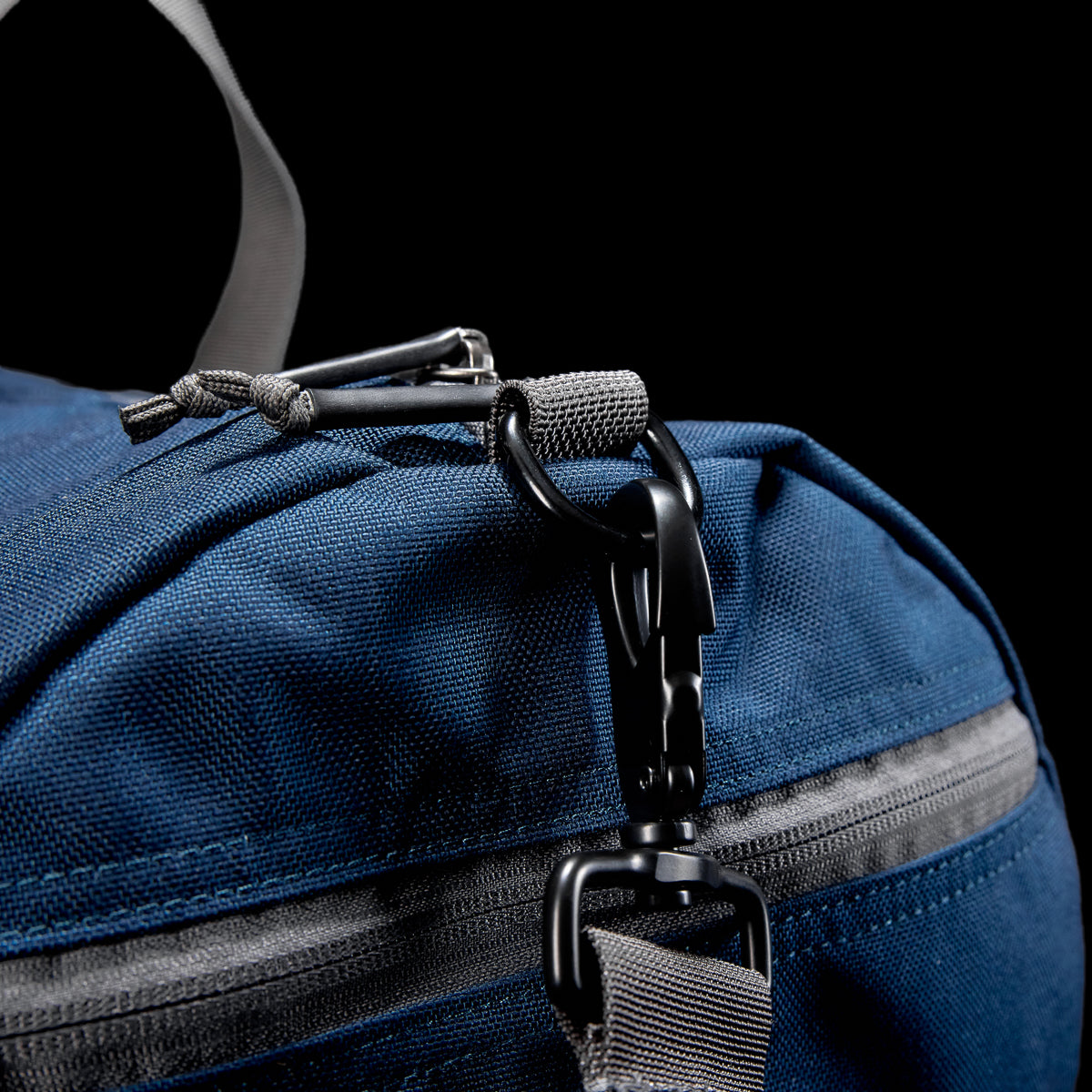 A close-up of a blue Gym Bag - Cordura made from durable fabric featuring a black carabiner on one loop, against a black backdrop.