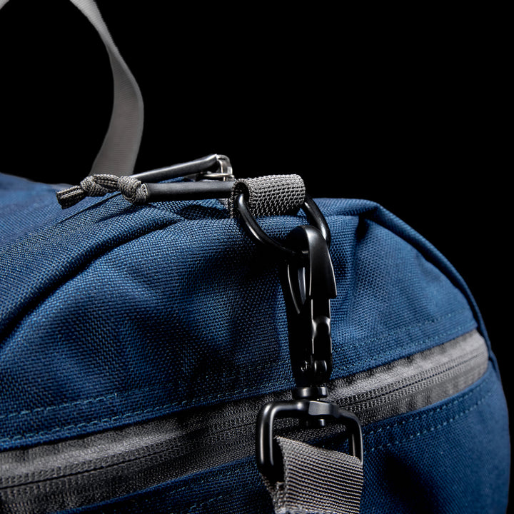A close-up of a blue Gym Bag - Cordura made from durable fabric featuring a black carabiner on one loop, against a black backdrop.