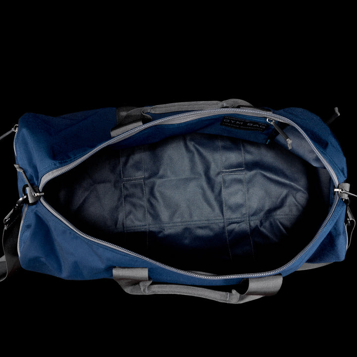 The Gym Bag - Cordura in navy blue, crafted from durable CORDURA fabric with gray handles, is displayed from above on a sleek black background.