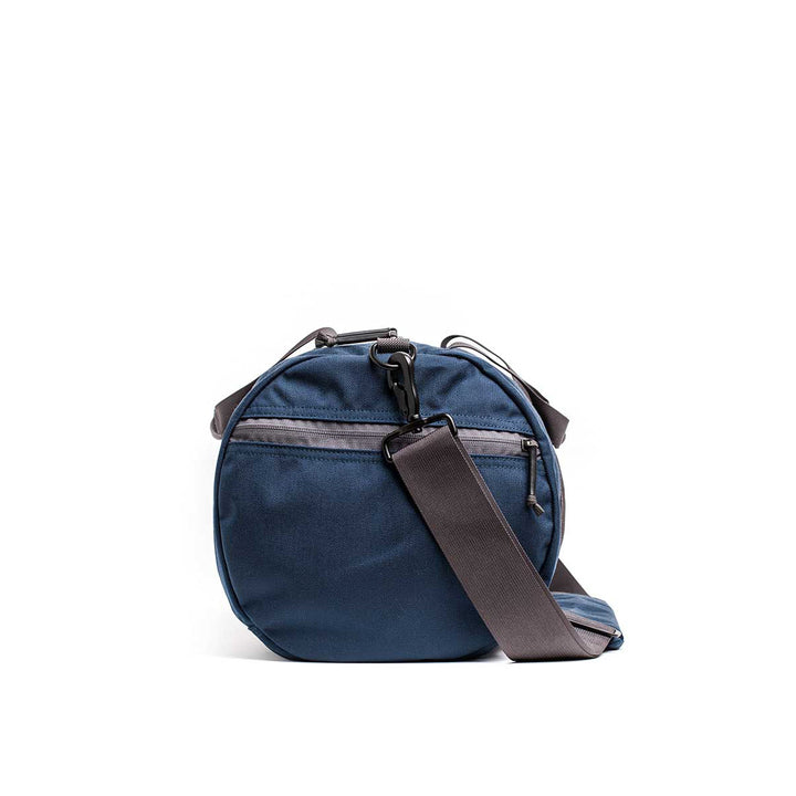 The Gym Bag - Cordura is a round blue shoulder bag made from durable CORDURA fabric, featuring zip pockets and an adjustable black strap. It's the ideal gym companion for style and reliability, set against a white background.