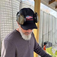 Man with white beard wearing black GORUCK cap and ear protection headphones at outdoor shooting range