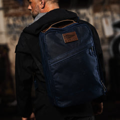 A person stands in an urban setting, wearing a black jacket and carrying a Heritage Rucksack, the navy GR1 USA - Heritage backpack crafted from Waxed Duck Canvas with brown detailing for enduring style.