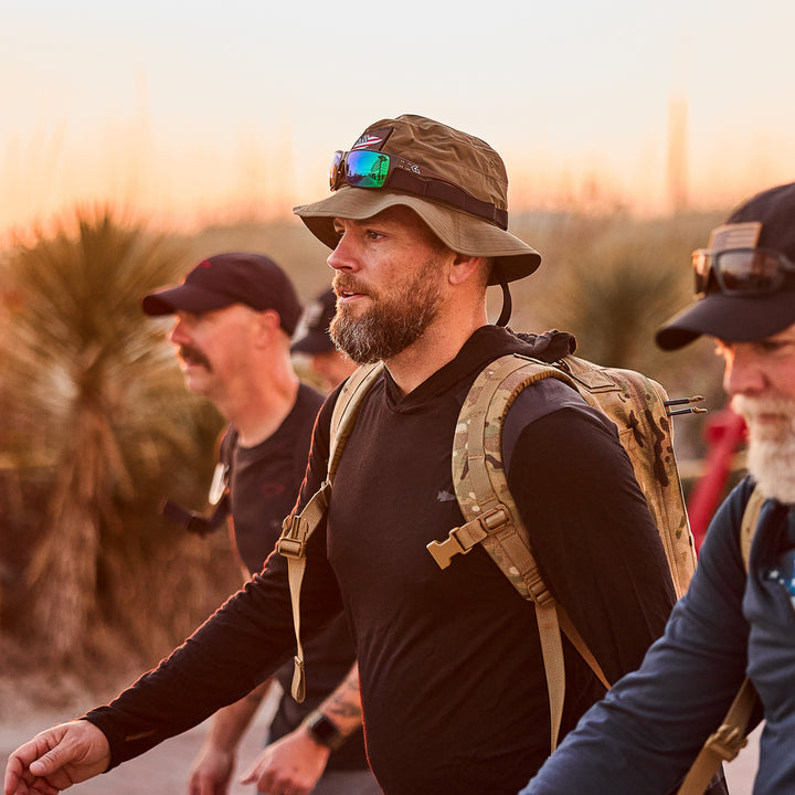 Three men hiking at sunset wear military-style gear with backpacks and Boonie Hats made of ToughDry® fabric, set against a blurred tree and sky background.