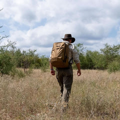 Person hiking through dry grassland wearing a brown backpack and wide-brim hat under cloudy sky