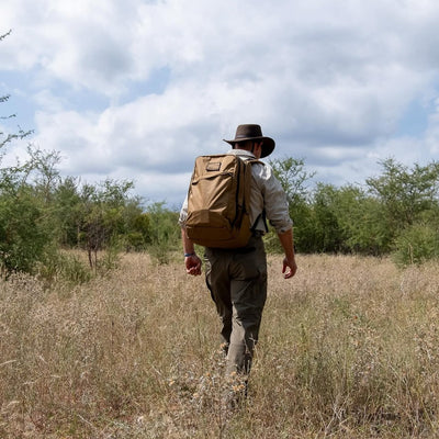 With the GR2 X-PAC backpack and a hat, a traveler strides through tall grass under a cloudy sky—ready for adventure with durable gear.