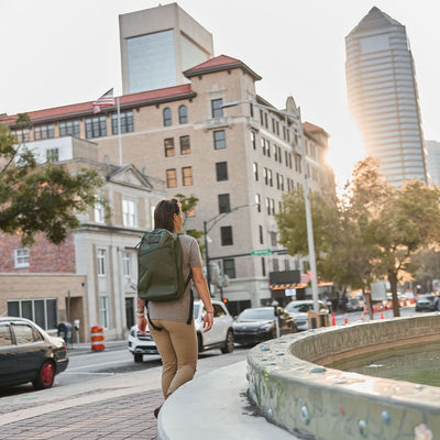 A person carrying the Bullet X-PAC daypack with AquaGuard zippers walks past a city fountain, tall buildings and cars in the background beneath a sunset sky.