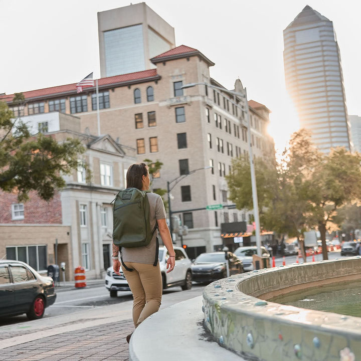 A person carrying the Bullet X-PAC daypack with AquaGuard zippers walks past a city fountain, tall buildings and cars in the background beneath a sunset sky.
