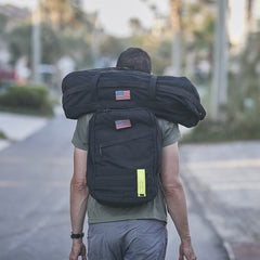 A person with a black backpack and a Sandbag Kit (w/ Filler Bag), both featuring US flag patches, walks down a street.