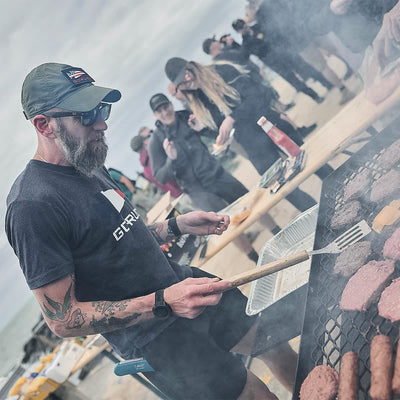 A person wearing a GORUCK Performance TAC Hat made from TOUGHDRY sweat-wicking material grills burgers and cheese on a beach. Nearby, people chat and enjoy their food. The overcast sky adds to the casual and relaxed atmosphere, emphasizing the beachside barbecue gathering.
