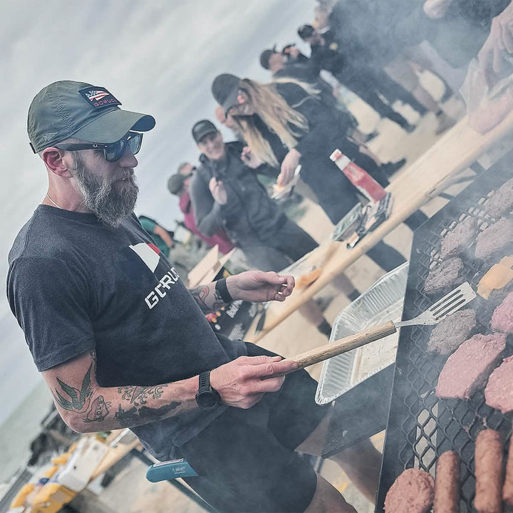 A person wearing a GORUCK Performance TAC Hat made from TOUGHDRY sweat-wicking material grills burgers and cheese on a beach. Nearby, people chat and enjoy their food. The overcast sky adds to the casual and relaxed atmosphere, emphasizing the beachside barbecue gathering.