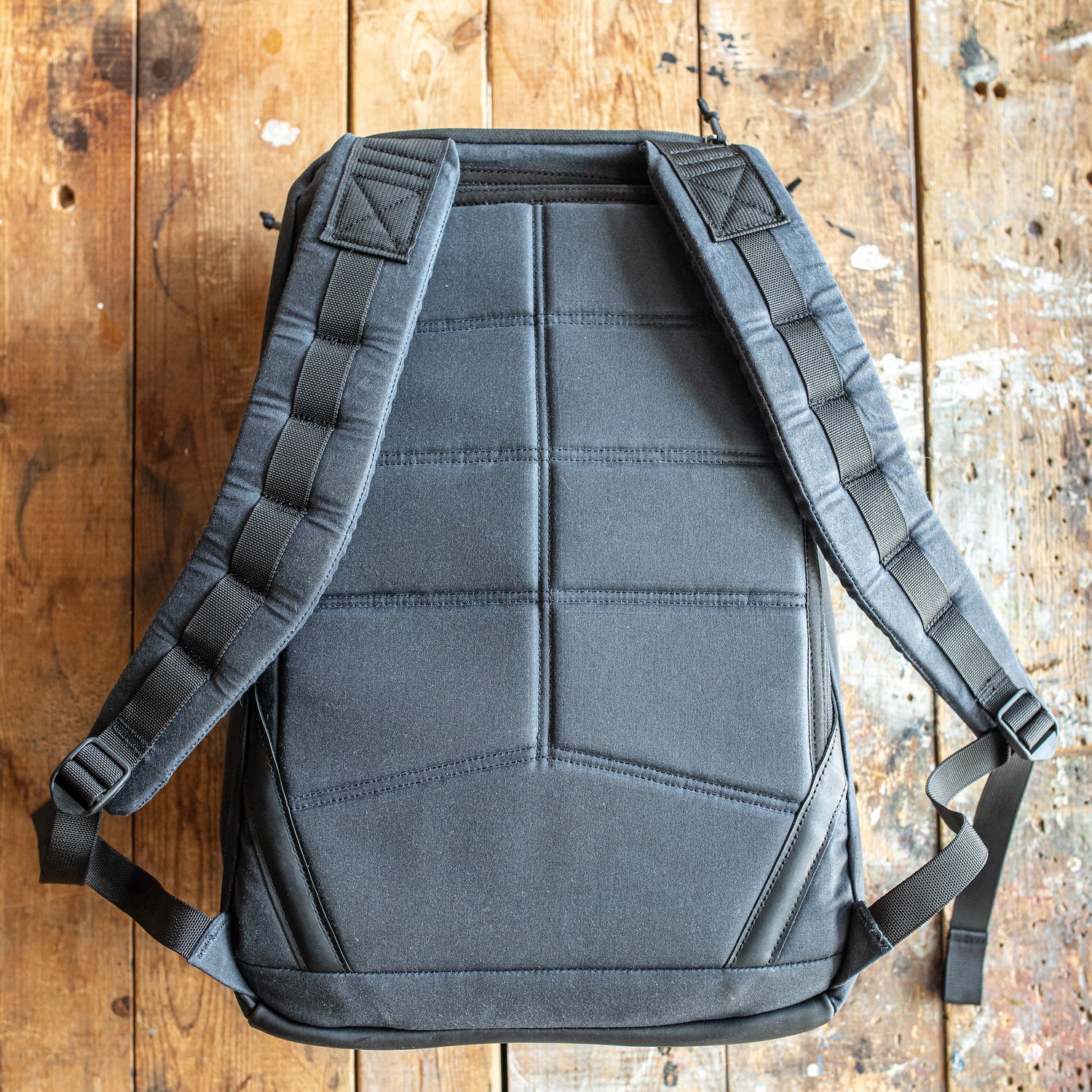A GR1 USA - Heritage Waxed Canvas rucksack in black with padded straps lies flat on a worn wooden surface.