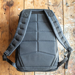 A GR1 USA - Heritage Waxed Canvas rucksack in black with padded straps lies flat on a worn wooden surface.