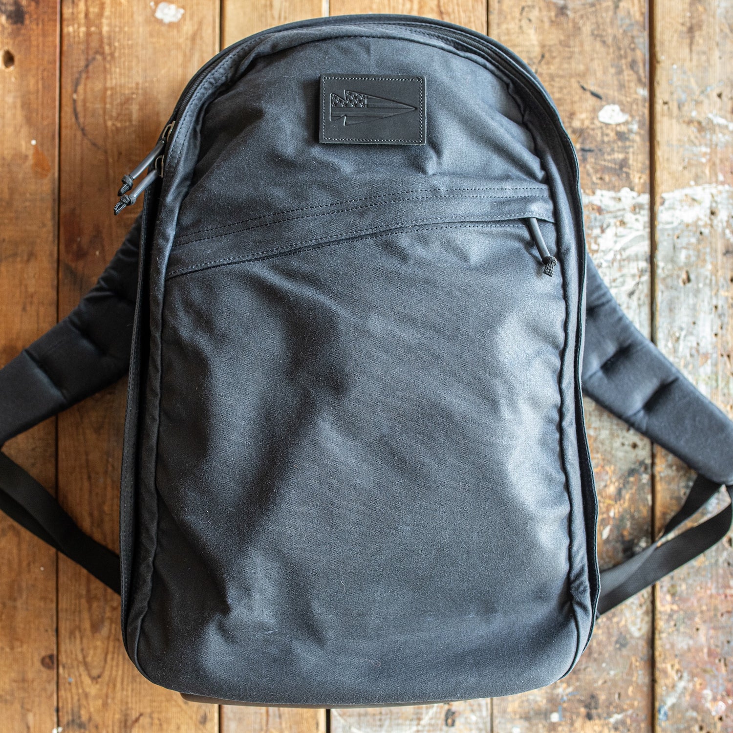 A GR1 USA - Heritage Waxed Canvas backpack in black rests on a worn wooden surface, featuring a convenient front zipper pocket.