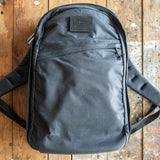 A GR1 USA - Heritage Waxed Canvas backpack in black rests on a worn wooden surface, featuring a convenient front zipper pocket.