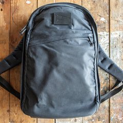 A GR1 USA - Heritage Waxed Canvas backpack in black rests on a worn wooden surface, featuring a convenient front zipper pocket.