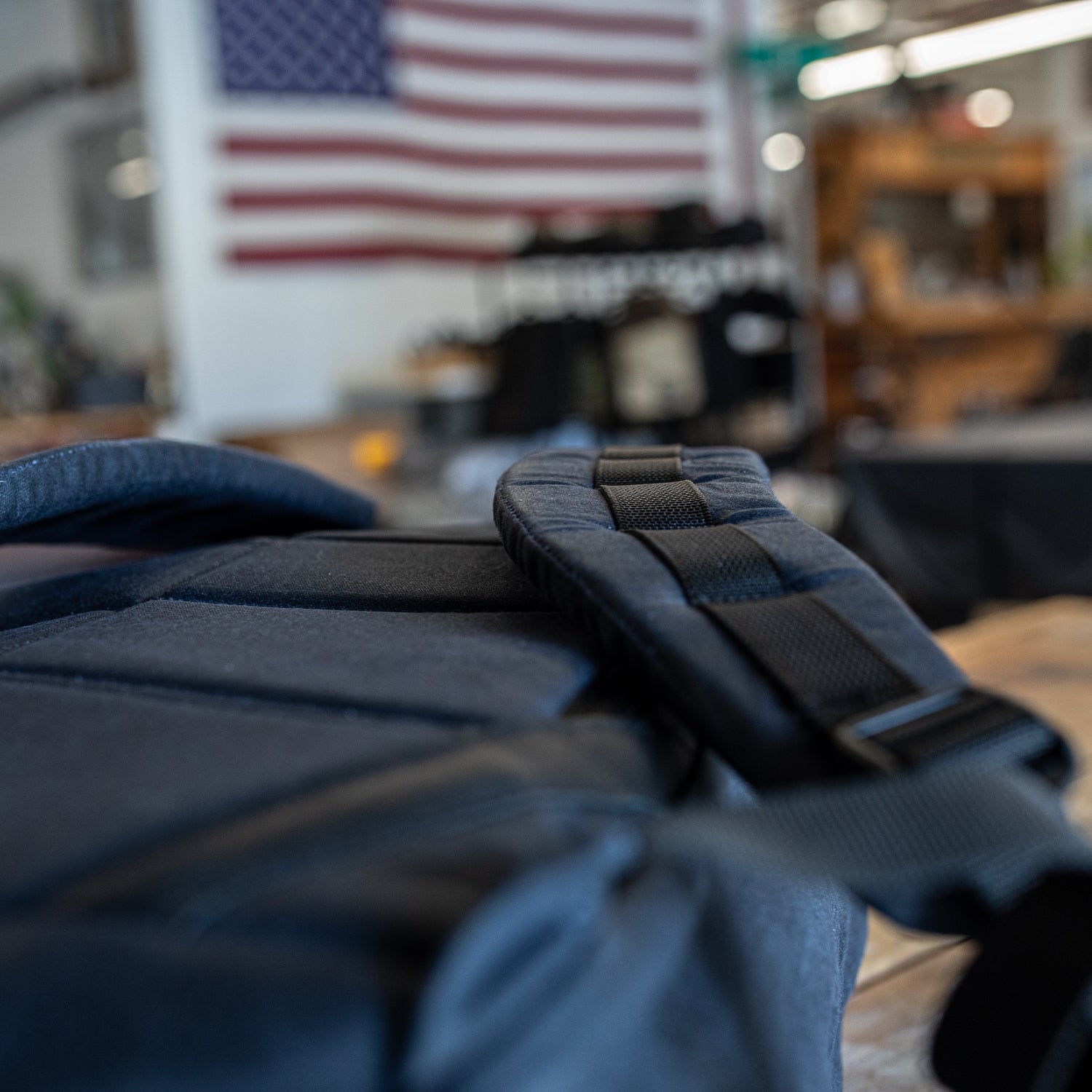 Close-up of a black strap from the GR1 USA - Heritage Waxed Canvas rucksack, with a blurred American flag hanging in the background.