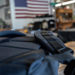 Close-up of a black strap from the GR1 USA - Heritage Waxed Canvas rucksack, with a blurred American flag hanging in the background.