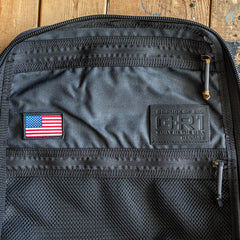 Open GR1 USA - Heritage Waxed Canvas rucksack in black, featuring a mesh pocket, USA flag patch, and an embossed “GORUCK” label, displayed on a wooden surface; expertly crafted from durable waxed duck canvas for a timeless, heritage-inspired appeal.