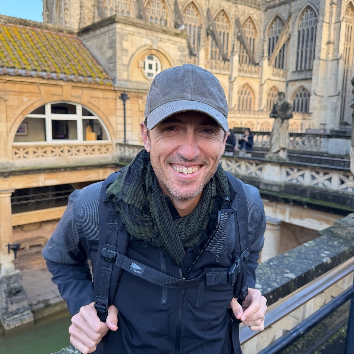 A smiling man wears the Tac Hat - Heritage as he stands outside near historic stone architecture and water.