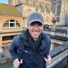 A smiling man wears the Tac Hat - Heritage as he stands outside near historic stone architecture and water.