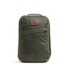 The GR1 USA - Heritage Waxed Canvas rucksack in olive green features a brown Red Wing Leather patch and a front diagonal zipper pocket, shown standing upright on a white background.