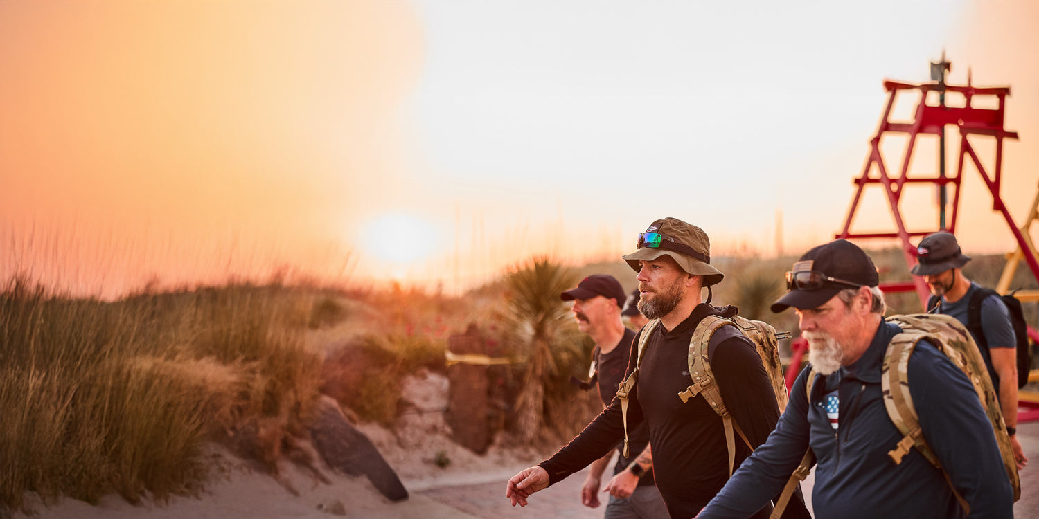 Four men with backpacks walk along a sandy path at sunset, with tall grass and a lifeguard tower nearby.