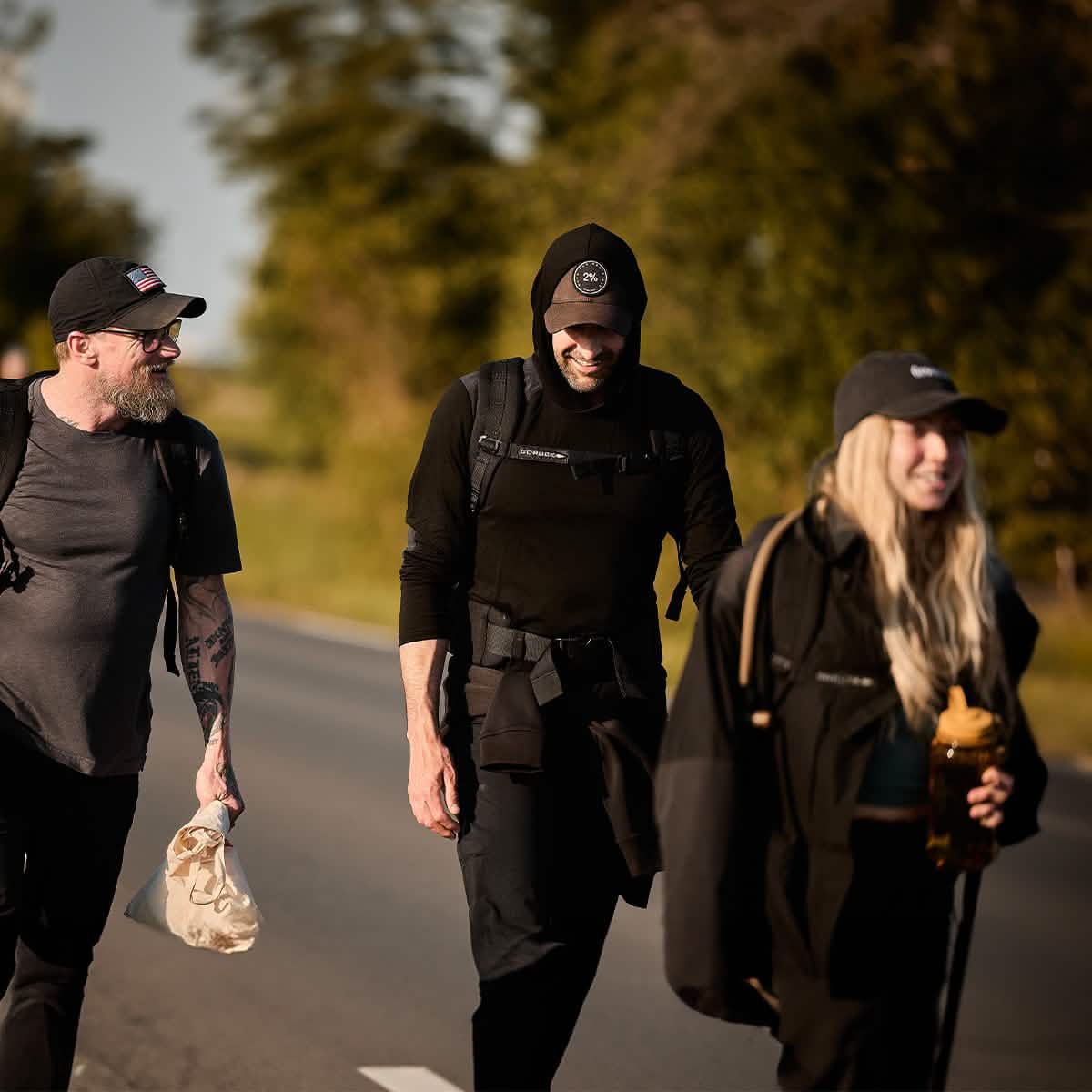 Three people stroll along a road, clad in black attire and hats reminiscent of the Men’s Commando Pullover - Merino Wool by GORUCK. The outdoor scene is framed by trees and bushes, with one person holding a cup and another carrying a smaller bag. They seem to be enjoying the sunny day.