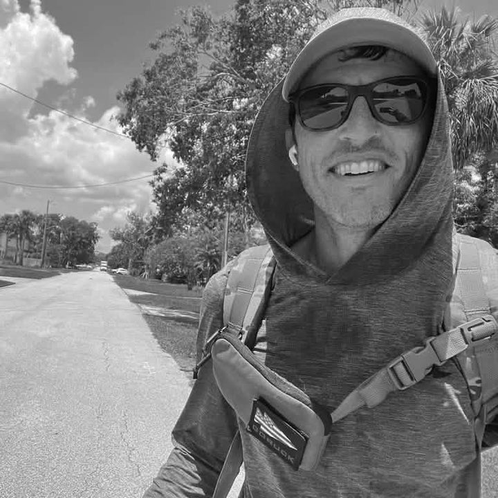 A person wearing sunglasses, a hat, and a hooded jacket smiles at the camera. They have a backpack and a sleek Sternum Pouch - Cordura from GORUCK. The background shows a sunny street lined with trees and a partly cloudy sky.
