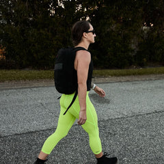 Person walking outdoors wearing neon yellow leggings, black tank top, sunglasses, and a black backpack