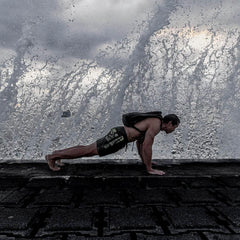 Man doing a push-up with a backpack on a wet stone surface as large ocean waves crash behind him