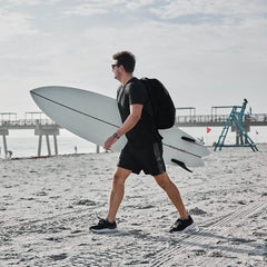 Man walking on beach carrying white surfboard wearing black athletic shorts and backpack in coastal setting