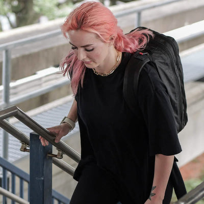 A person with pink hair is ascending outdoor stairs while holding a GORUCK Bullet Ruck Double Compartment made from durable Dyneema. They're dressed in a black shirt and a gold chain necklace, set against a backdrop of concrete structures and greenery that accentuates the chic mix of urban and natural elements.