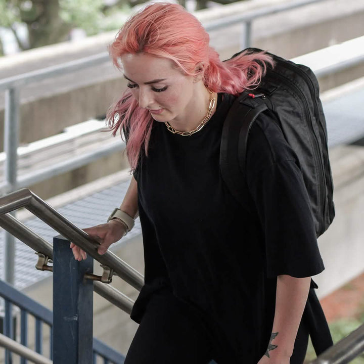 A person with pink hair is ascending outdoor stairs while holding a GORUCK Bullet Ruck Double Compartment made from durable Dyneema. They're dressed in a black shirt and a gold chain necklace, set against a backdrop of concrete structures and greenery that accentuates the chic mix of urban and natural elements.