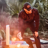 Man in black GORUCK fleece hoodie and hat grabs a drink from cooler outdoors near campfire