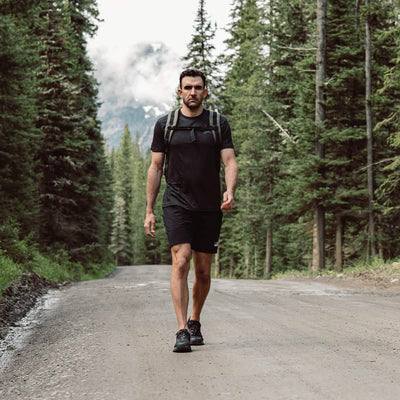 A man walks confidently down a dirt road surrounded by tall, dense trees. He's wearing a black T-shirt, shorts, hiking shoes, and ankle Merino Challenge Socks from GORUCK with a backpack. Misty mountains are visible in the background under a cloudy sky.