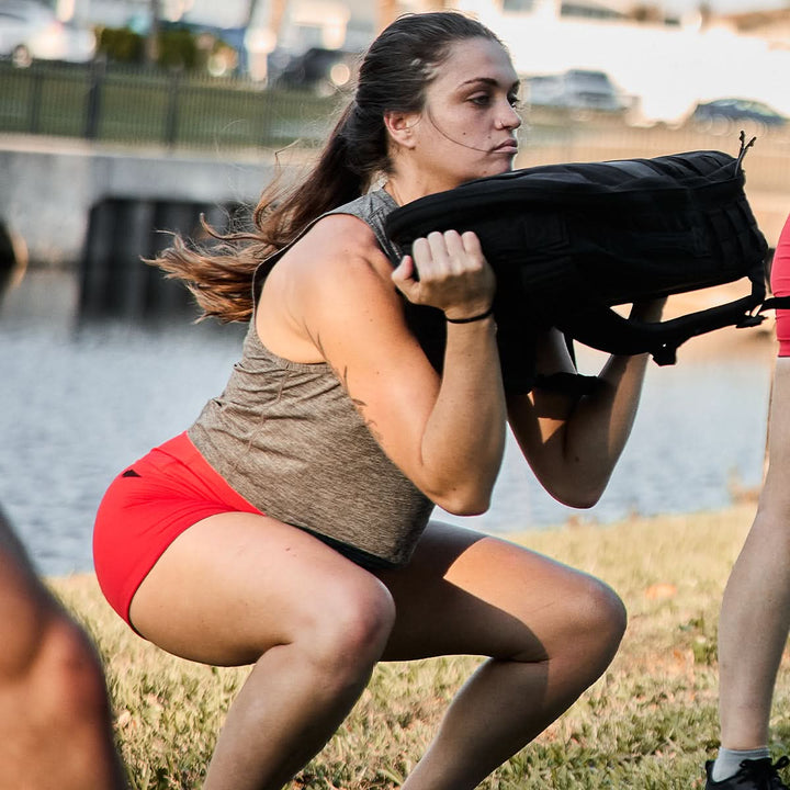 A woman performs squats outside with a weighted bag, highlighting her red Women’s Squat Shorts - ToughFlex from GORUCK. The shorts incorporate ToughFlex Fabric and no-ride technology, which she complements with a gray tank top for an active ensemble.